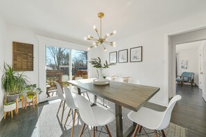 Un comedor con una mesa de madera, sillas blancas, una araña y cuadros en la pared.