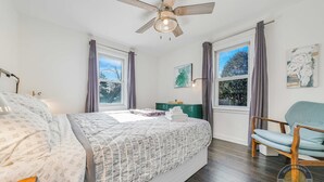Ein Schlafzimmer mit einem Bett, einem Sessel, einem Deckenventilator und zwei Fenstern mit Vorhängen.