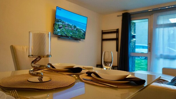 A dining area with a table set for two, a television mounted on the wall, and a view of the outdoors through a window. - Stunning 1-bed Apartment in Bishop Auckland (Bishop Auckland)