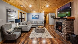A cozy living room with a stone fireplace, a large flat-screen TV, a gray sectional sofa, and a patterned armchair. - Serene Cabin, Sleeps 6・Hot Tub・Pool Table & Arcade (Pigeon Forge)