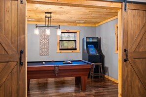 A pool table with a billiard cue, a pool table, a window with blinds, a wall-mounted decorative panel, and a pool table with a ball rack.