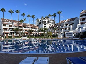 Ein Resort mit einem zentral gelegenen Pool, umgeben von Liegestühlen, Palmen und mehrstöckigen Gebäuden mit Balkonen.