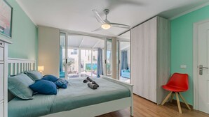 Une chambre à coucher avec un lit, un fauteuil rouge, un ventilateur de plafond et une vue sur un espace extérieur, visible à travers une porte vitrée.