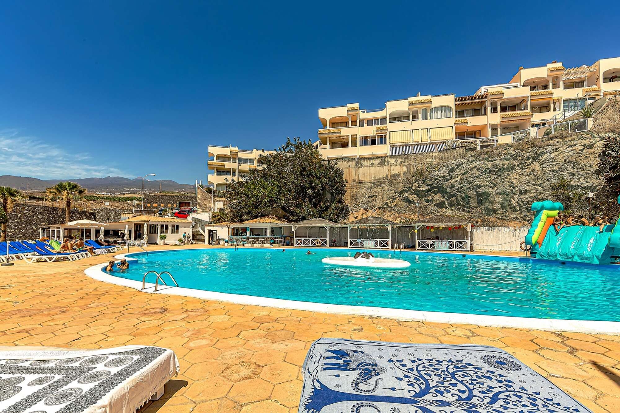 A swimming pool with lounge chairs and umbrellas, a lifeguard stand, and a building with balconies.