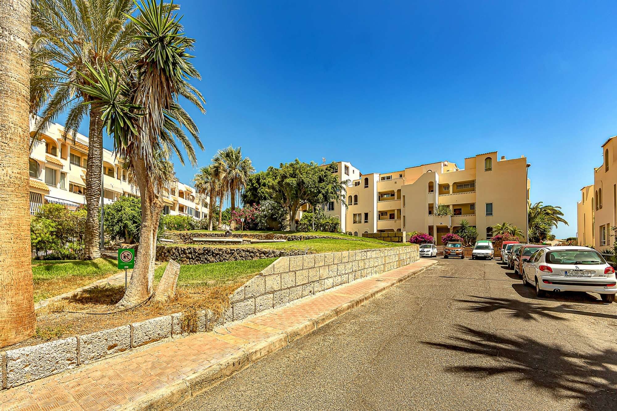 A residential street with multi-story buildings, a stone retaining wall, and parked cars.