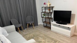 Un salon avec un canapé gris, deux fauteuils gris, un téléviseur posé sur un meuble blanc, un plancher en bois et un rideau gris.
