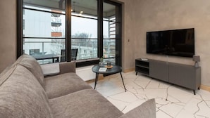 A modern living room with a large window, a flat-screen TV, a grey sofa, and a small round table.