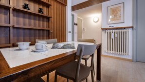Un comedor con una mesa de madera, sillas y suelo de baldosas blancas. Hay estantes con adornos y un cuadro en la pared.