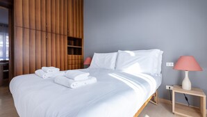 Una cama bien hecha con sábanas blancas y toallas dobladas, un armario de madera, una mesita de noche con lámpara y un pequeño estante.