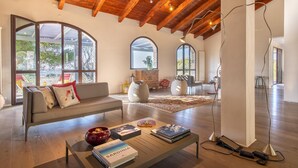 A spacious living room with a high wooden ceiling, large arched windows, a sofa, a coffee table with books, and a modern lamp. - Sardinia Re - Villa Posta Del Sol (Alghero)