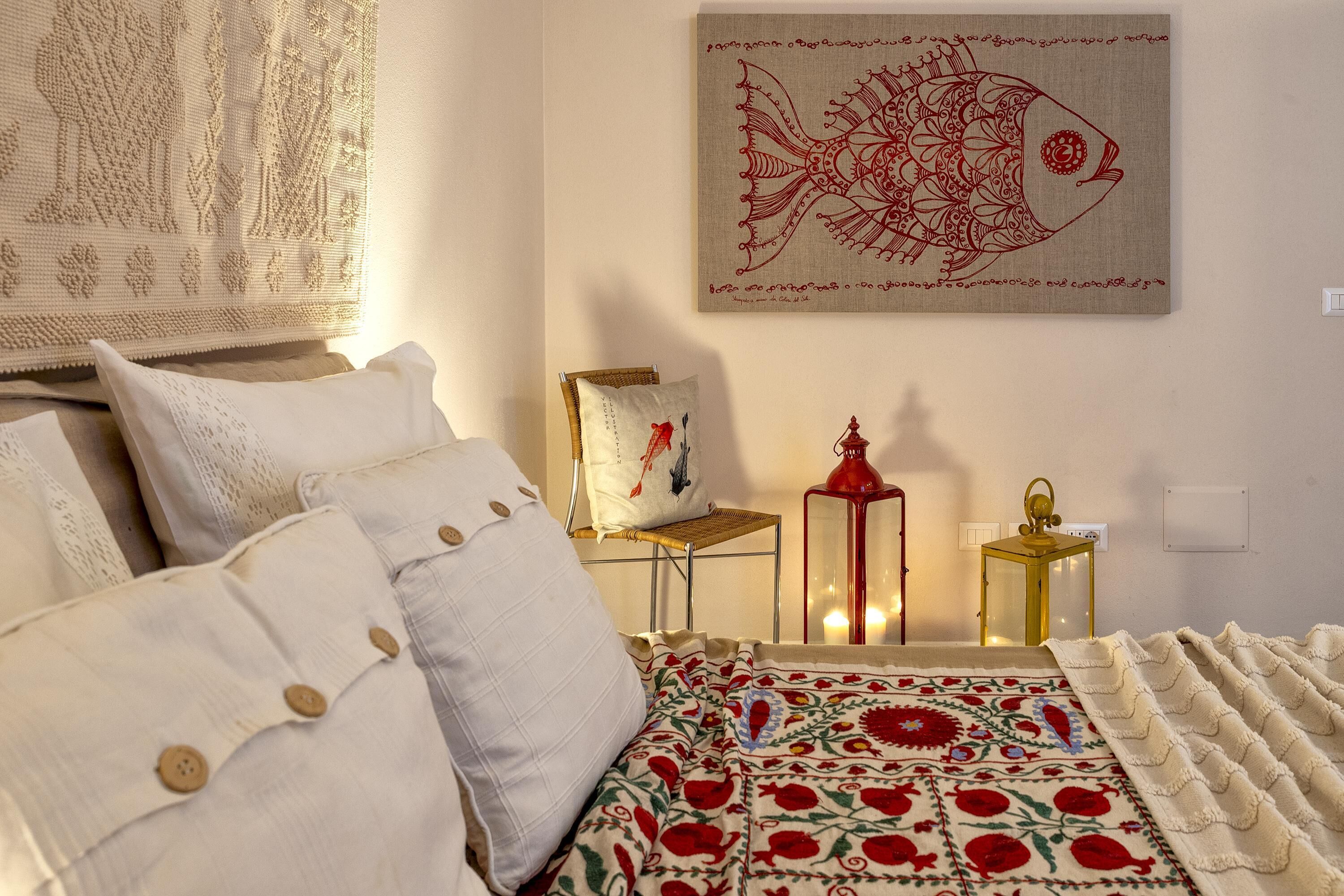 Een slaapkamer met een bed, kussens, een deken, een wanddecoratie, een stoel en een tafeltje met een lamp.