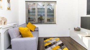 A modern living room with a grey sofa, yellow cushions, a black and yellow rug, a large window with blinds, and a flat-screen TV. - Stunning 1-bed Apartment in Birmingham (Birmingham)