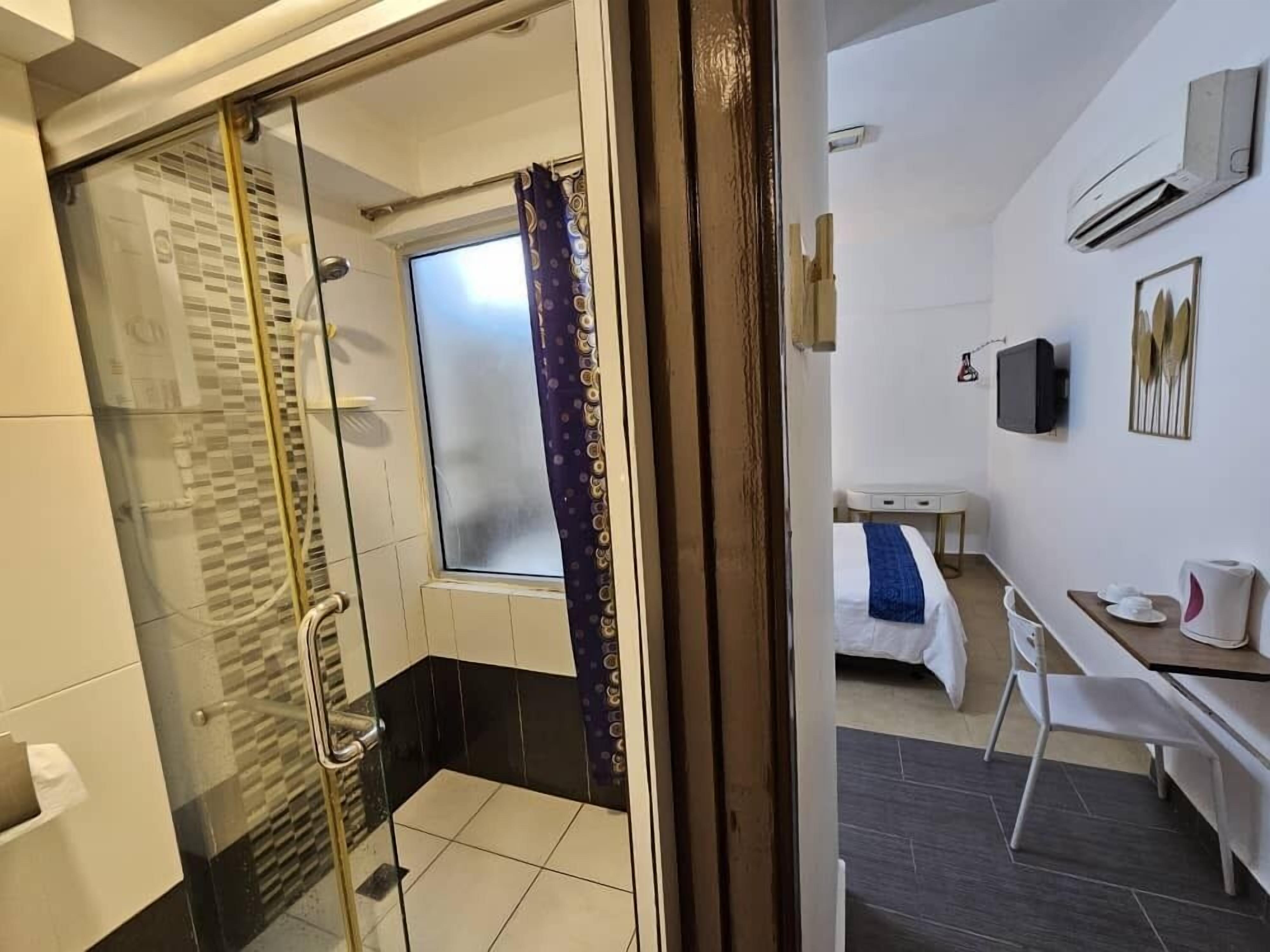 A hotel room with a glass shower enclosure, a bed with a blue bedspread, a desk with a chair, and a TV mounted on the wall.