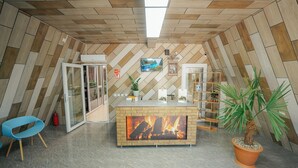 Un área de recepción moderna con techo de madera en patrón de espiga, una chimenea central, una silla azul y una palma en maceta.
