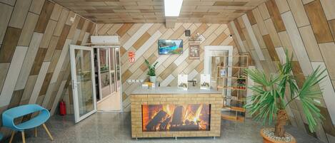 A modern reception area with a herringbone wood ceiling, a central fireplace, a blue chair, and a potted palm tree.