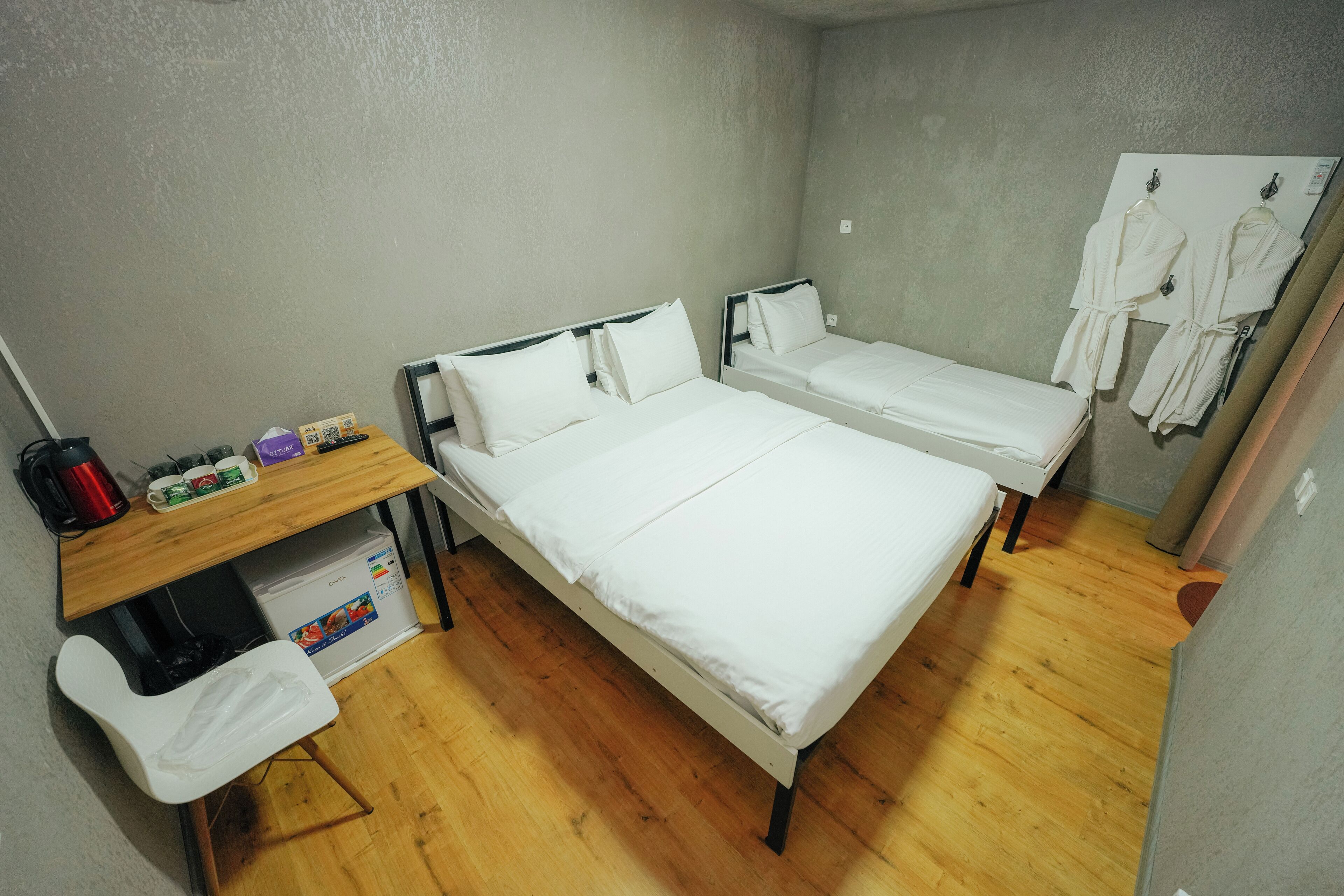 A room with two single beds, a wooden desk with a kettle, and a small refrigerator.