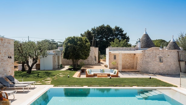 A poolside area with a stone building, a grassy lawn, and a seating area with a sofa and cushions. - Donna Vincenza in Selva di Fasano (Monopoli)
