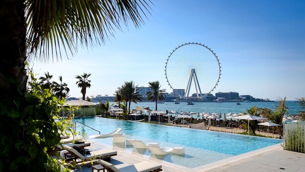 A poolside area with lounge chairs, a Ferris wheel in the background, and a view of the sea. - Elite 2BR at La Vie with sea view (Dubai)