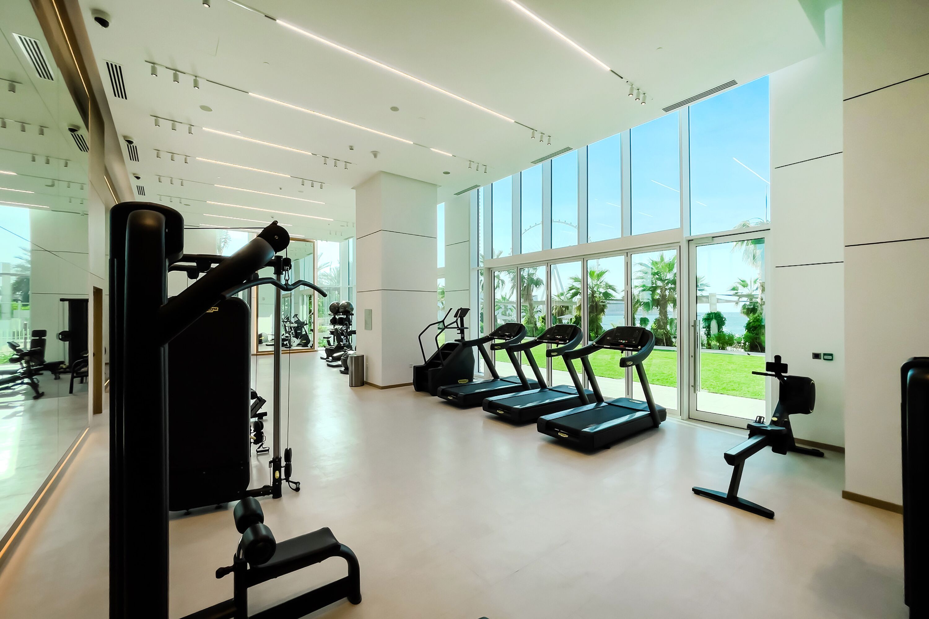 A modern gym with treadmills, weight machines, and large windows offering views of greenery.