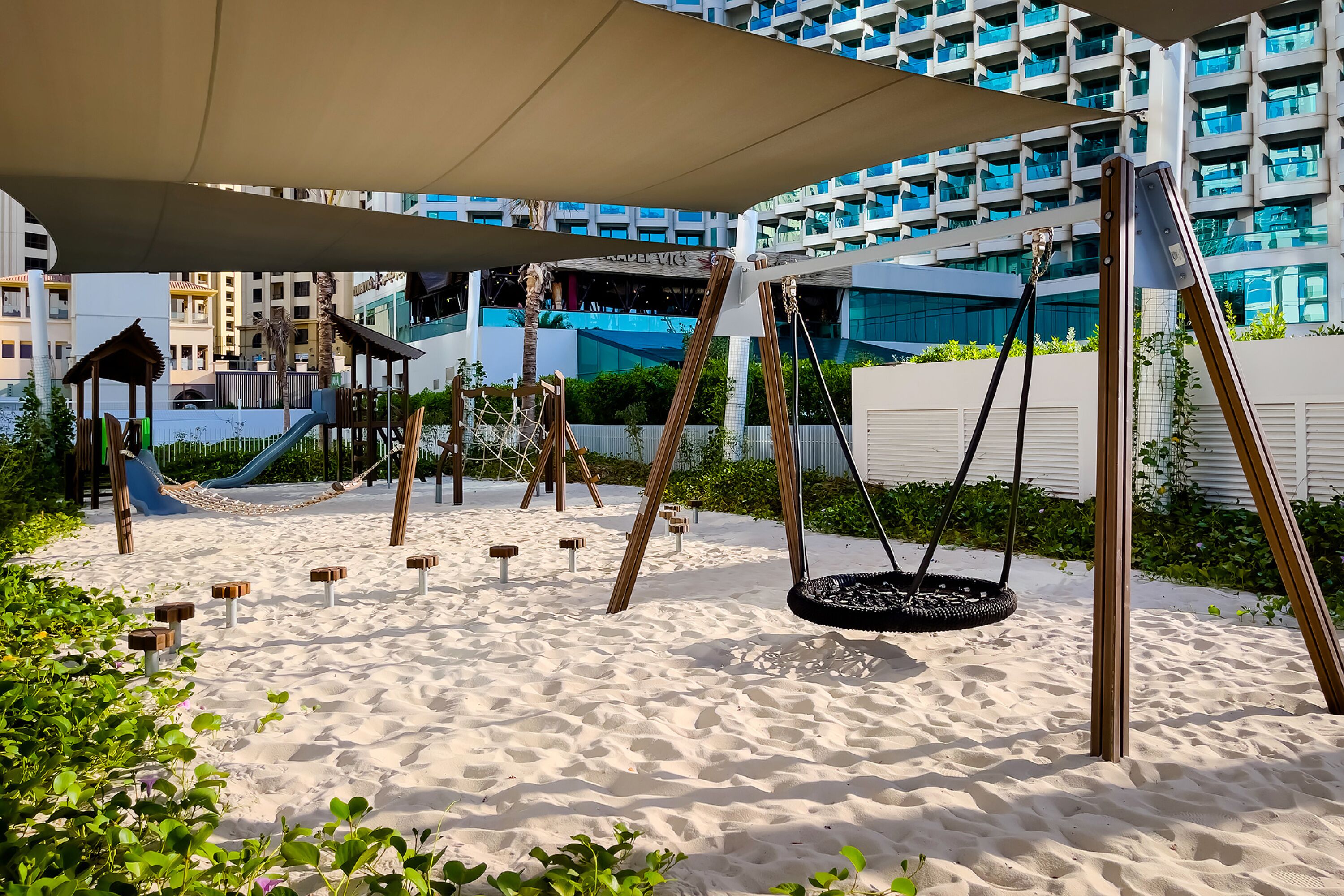 A playground with swings and a slide, surrounded by sand and greenery, with modern buildings in the background.