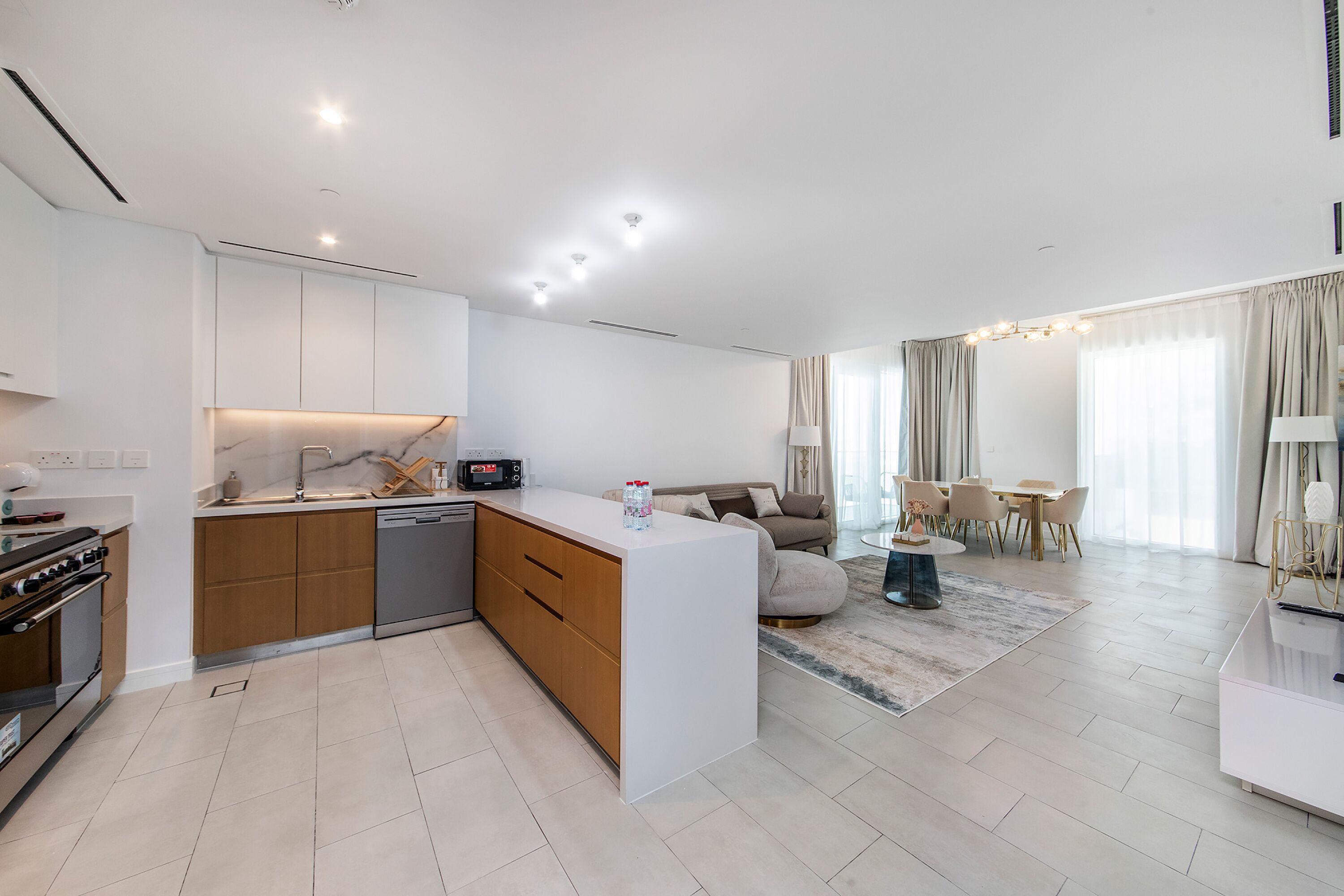 A modern kitchen with a central island, built-in oven, and dishwasher. There is a dining area with a table and chairs, and a living room with a sofa and a coffee table.