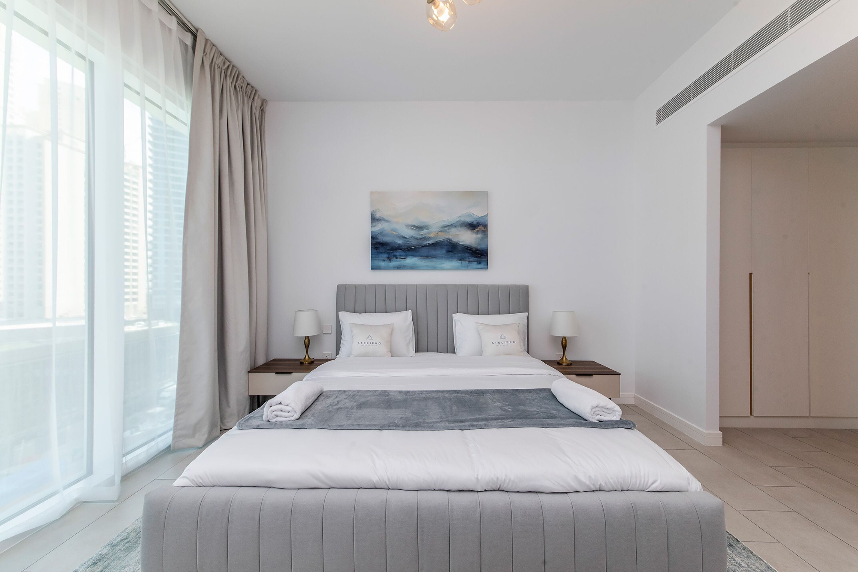 A modern hotel room with a large bed, a painting above it, and a view of the city through the window.