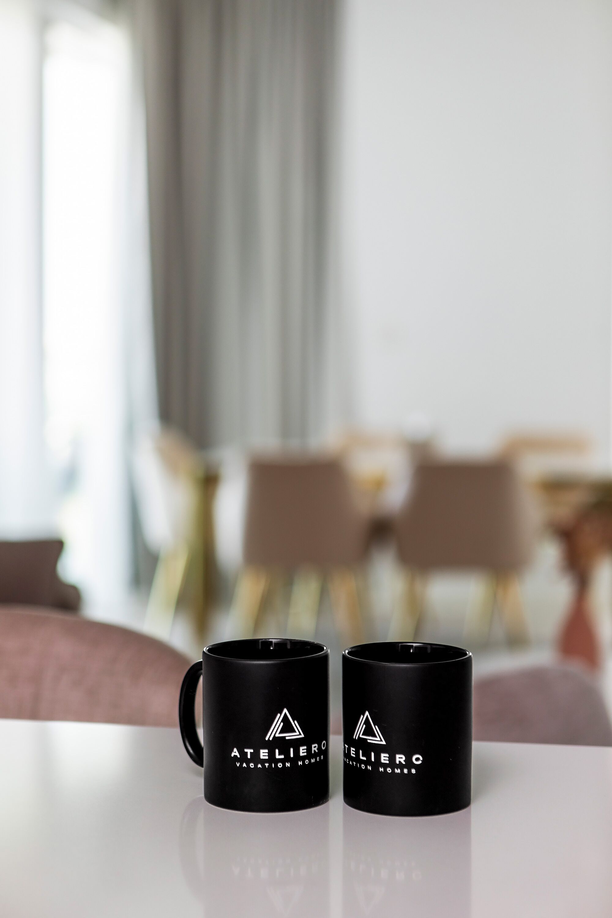 Two black mugs with a logo on a reflective surface in a room with a blurred background.