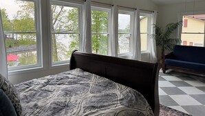 A bedroom with a large bed, multiple windows, a view of a lake, a blue sofa, and a potted plant.