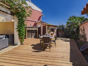 Une terrasse en bois avec une table et des chaises, entourĂ©e de verdure et bordĂ©e dâun bĂątiment Ă la façade rouge.