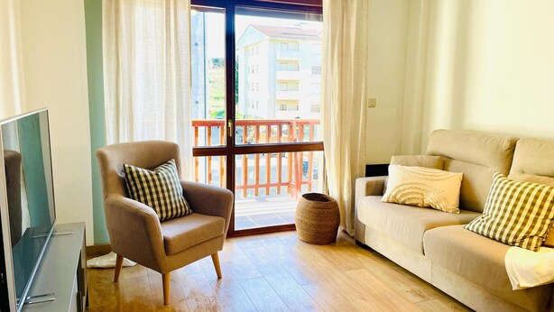 A living room with a sofa, armchair, TV, and a view of a balcony with buildings in the background. - Two-Bedroom Apartment with Pool, Balcony, and Garage in Tranquil Jaca (Jaca)