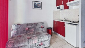 A compact kitchen with a red and white color scheme, a sofa with a London-themed print, a microwave, and a small trash bin. - Stylish 2-Room Apartment with Balcony, Private Parking, Sleeps 4-6 (Marseillan-Plage)