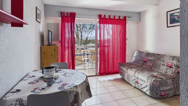 A room with a dining table, a sofa, a TV, and a view through a glass door. - Stylish 2-Room Apartment with Balcony, Private Parking, Sleeps 4-6 (Marseillan-Plage)