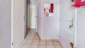 A hallway with a tiled floor, a white door, a mirror on the wall, and a red shelf. - Stylish 2-Room Apartment with Balcony, Private Parking, Sleeps 4-6 (Marseillan-Plage)