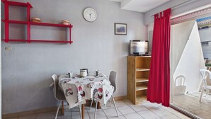 A dining area with a table set for two, a television on a wooden stand, a red curtain, and a window with a view. - Stylish 2-Room Apartment with Balcony, Private Parking, Sleeps 4-6 (Marseillan-Plage)