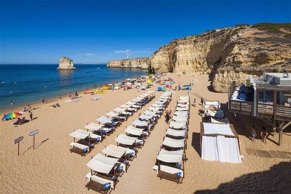 A beach with rows of sunbeds and umbrellas, a rocky cliff, and a lifeguard tower.