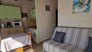 A small, well-lit room with a bed, a sofa, a kitchenette, and a painting on the wall.