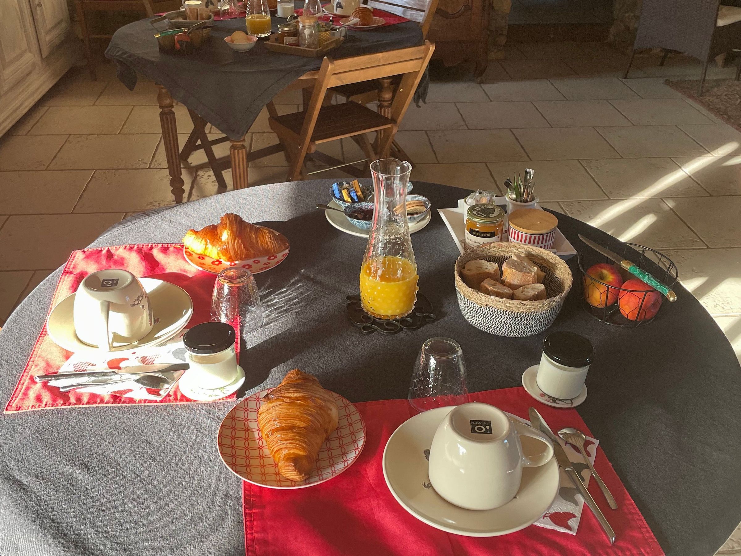 Une table de petit-déjeuner dressée avec des croissants, une bouteille de jus d’orange et divers aliments pour le petit-déjeuner.