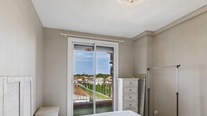 A bedroom with a bed, a dresser, a radiator, and a window with a view of a residential area. - Seaside Gem: Air-Conditioned 1-Bedroom Apartment with Ocean View Terrace at Grau d'Agde (Le Grau d'Agde)