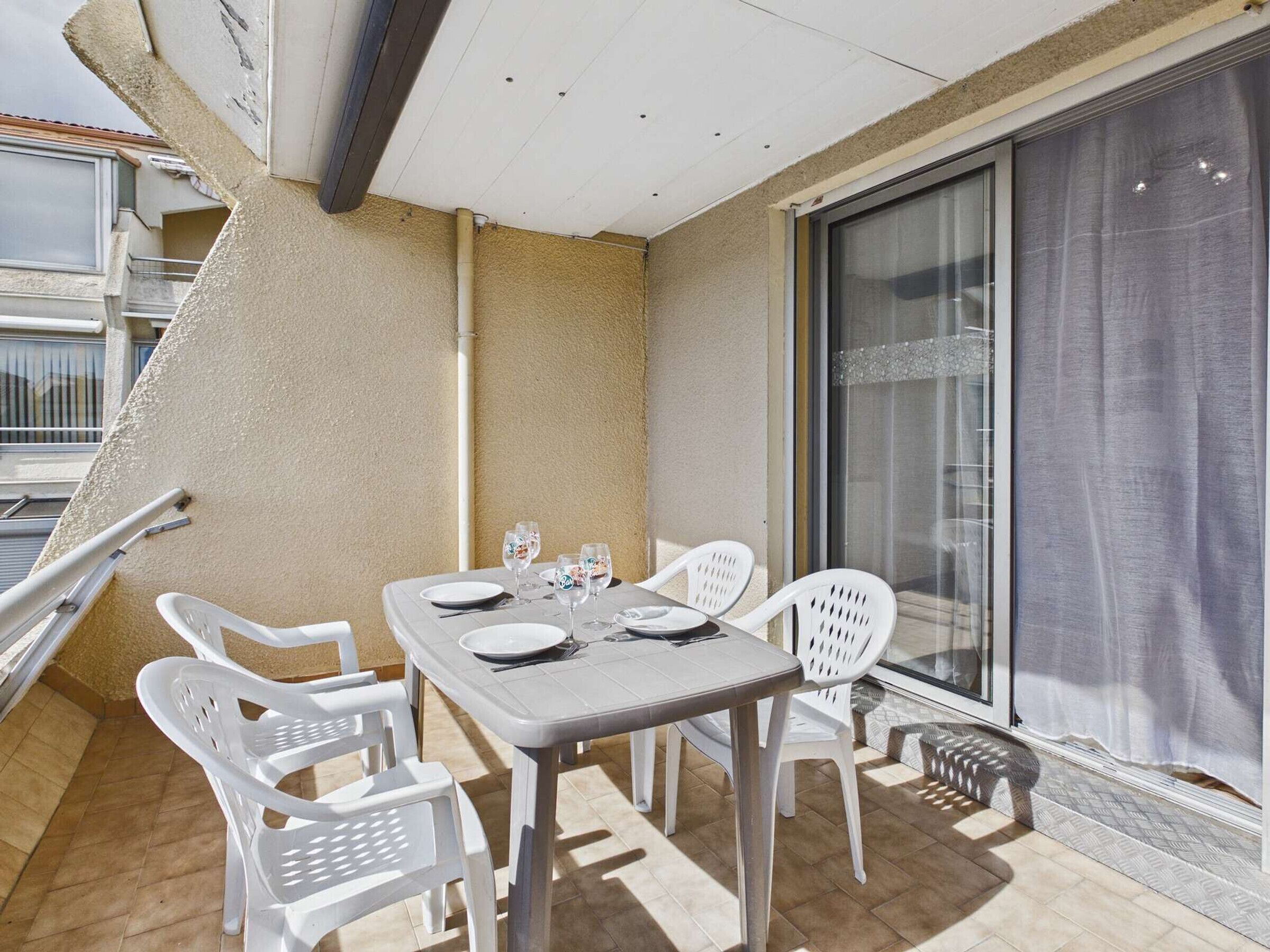 Un balcon avec une table dressée pour deux personnes, des chaises en plastique blanches et une porte vitrée donnant sur un espace abrité.
