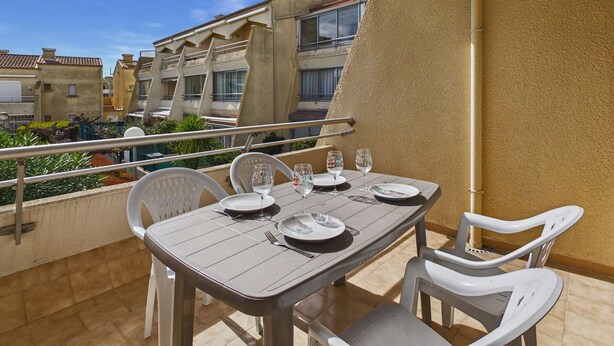 A balcony with a table set for two, chairs, and a view of neighboring buildings. - Cozy 2-Room Apartment for 4 with Pool View, Terrace, and Private Parking (Marseillan-Plage)