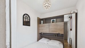 A bedroom with a bed, a wooden headboard, a small window, and a hallway leading to a door. - Cozy 2-Room Apartment for 4 with Pool View, Terrace, and Private Parking (Marseillan-Plage)