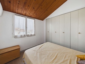 Une chambre avec un plafond en bois, une fenêtre avec des rideaux, un lit recouvert d’une couvre-lit à motifs et une armoire à plusieurs portes.