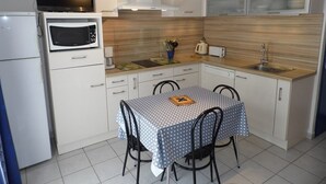 A compact kitchen with a microwave, stove, and sink. There is a dining table with chairs and a small TV mounted above it. - Modern 2-Bedroom Townhouse with Terrace, Private Parking & Pool Access in Marseillan (Marseillan-Plage)