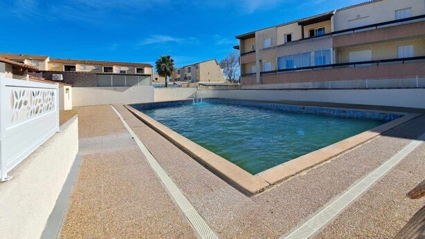 A swimming pool with a clear blue sky, a white fence, and buildings in the background. - Modern 2-Bedroom Townhouse with Terrace, Private Parking & Pool Access in Marseillan (Marseillan-Plage)