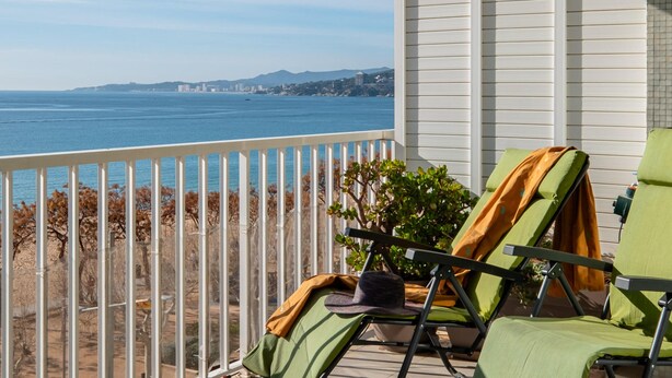 Un balcon avec deux fauteuils de jardin verts, un pot de fleurs et une vue sur l’océan et le paysage urbain lointain.