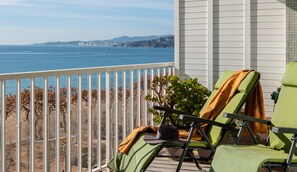 A balcony with two green lounge chairs, a potted plant, and a view of the ocean and distant cityscape. - Chic Palamós Seafront Apartment (Palamós)
