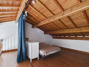 Un dormitorio ático pequeño y de una sola habitación, con techo de madera inclinado, una cama, un aparador blanco y una ventana con cortinas azules.