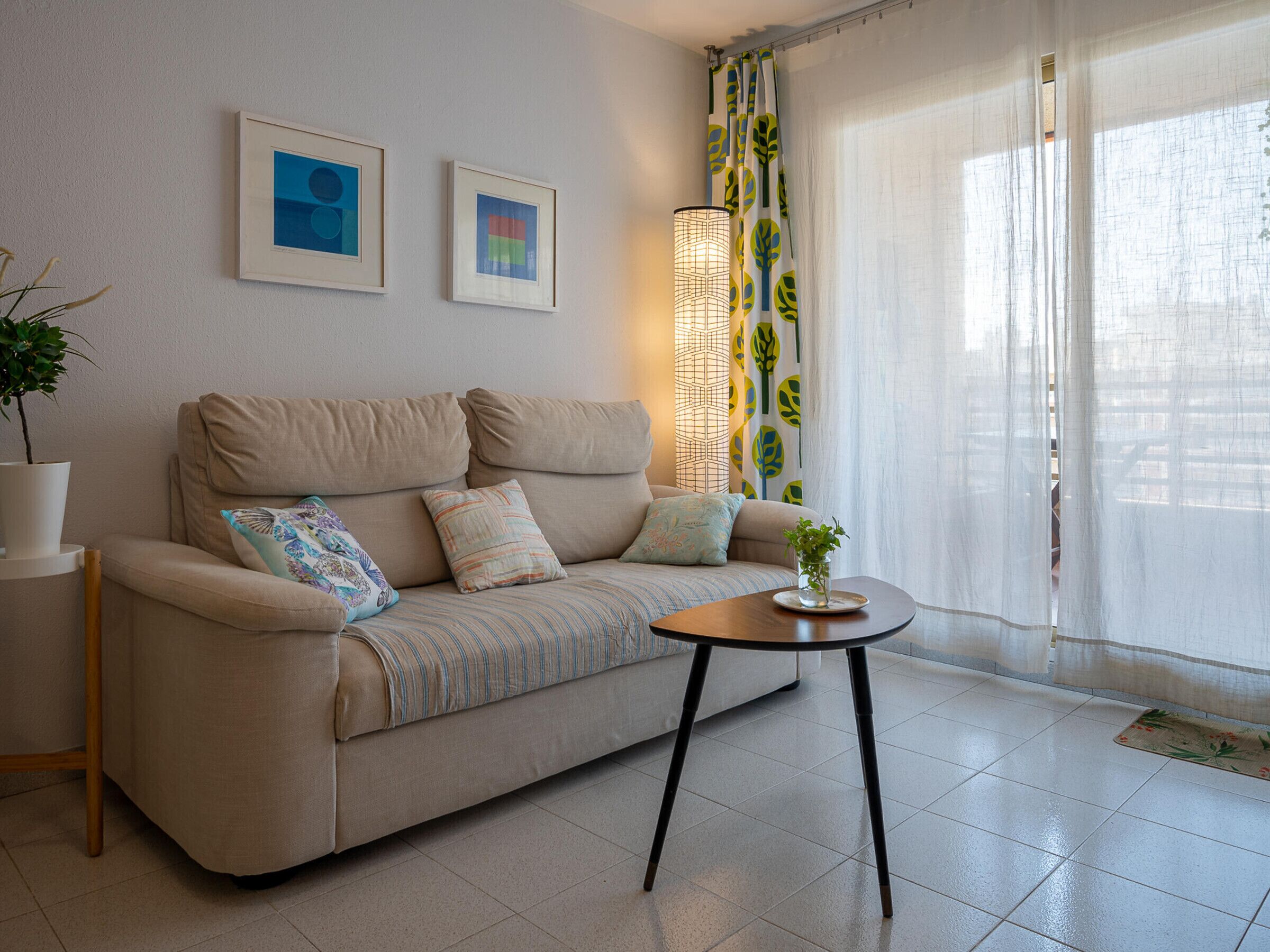 A modern living room with a beige sofa, two framed artworks, a floor lamp, and a view through sheer curtains.