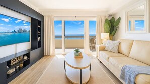 Una sala de estar moderna con un televisor grande, un sofá color beige, una mesa redonda y vistas al océano a través de ventanas amplias.