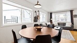 Amplio salón con una mesa redonda de madera, sillas negras, un sofá gris y un ventanal con vistas a una terraza y árboles.